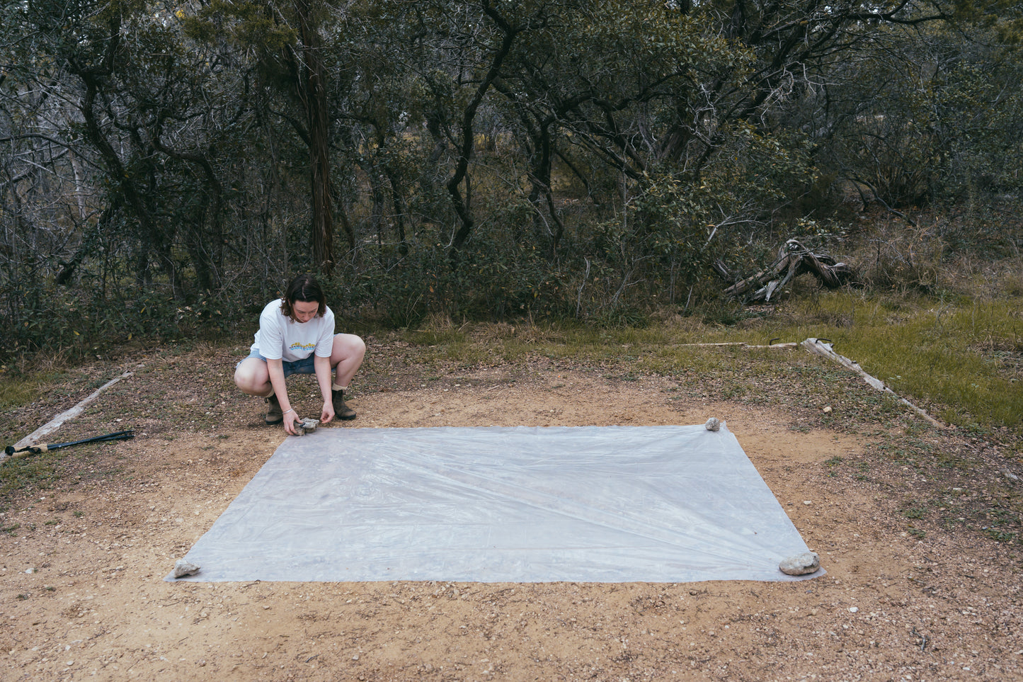 Person setting up a large gray mat on a dirt ground with trees in the background