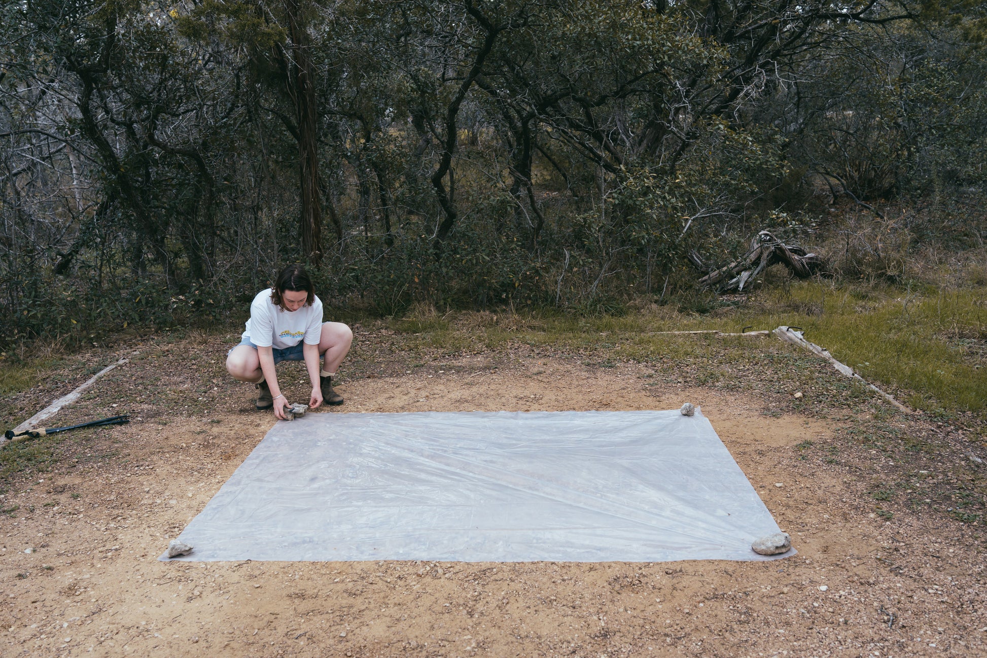 Person setting up a large gray mat on a dirt ground with trees in the background